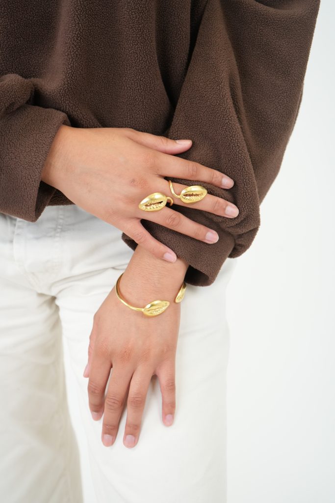 bague et bracelet Queen cauris portés par une femme en pantalon blanc et sweat marron.
